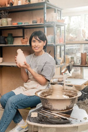 Lächelnde junge asiatische Kunsthandwerkerin in Schürze mit Keramikskulptur und Blick in die Kamera, während sie in der Nähe von feuchtem Ton auf Töpferscheibe in verschwommener Werkstatt sitzt, Konzept des Tonbildhauerprozesses