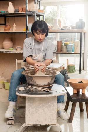 Téléchargez les photos : Jeune potier asiatique femelle en tablier moulant de l'argile humide sur roue de poterie près des outils et bol avec éponge tout en travaillant dans un atelier de céramique en arrière-plan, concept de processus de sculpture d'argile - en image libre de droit