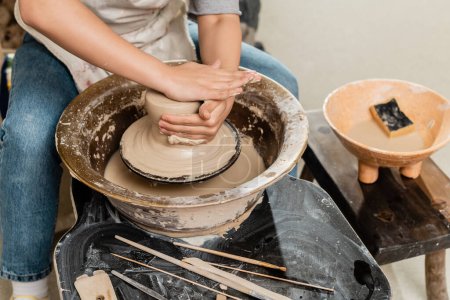 Téléchargez les photos : Vue grand angle de jeune artisan en tablier façonnant l'argile sur la roue de poterie et travaillant près des outils en bois et bol flou avec de l'eau dans un atelier de céramique, concept de processus de sculpture de l'argile - en image libre de droit