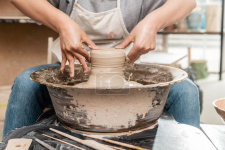 Ausgeschnittene Ansicht einer verschwommenen Kunsthandwerkerin in Schürze beim Formen von nassem Ton auf Töpferscheibe in der Nähe von Werkzeugen auf dem Tisch in der Keramik-Kunst-Werkstatt im Hintergrund, Tonbildhauerei-Prozess-Konzept