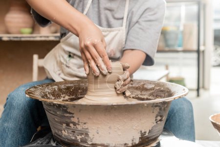Ausgeschnittene Ansicht einer verschwommenen jungen Kunsthandwerkerin in Schürze, die Ton auf Töpferscheibe gießt, während sie im Keramik-Atelier im Hintergrund arbeitet, Konzept des Tonbildhauerprozesses