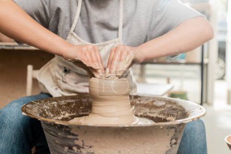 Téléchargez les photos : Vue recadrée du jeune potier femelle en tablier moulant de l'argile humide et travaillant avec une roue de poterie dans un atelier de céramique d'art flou en arrière-plan, concept de processus de sculpture d'argile - en image libre de droit
