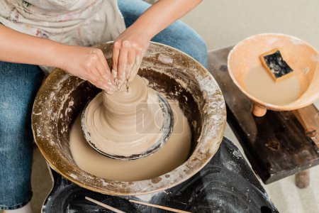High angle view of young female artisan in apron molding wet clay on pottery wheel and working near blurred bowl with water and sponge in ceramic studio, skilled pottery making concept