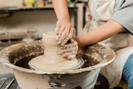 Vue recadrée de céramiste féminine floue dans le moulage de tablier d'argile humide et de travail sur la rotation de la roue de poterie en atelier de céramique d'art, concept de fabrication de poterie qualifiée