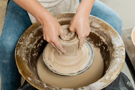 Vue grand angle du jeune potier femelle en tablier façonnant de l'argile humide tout en travaillant sur la rotation de la roue de poterie dans un atelier d'art en arrière-plan, concept de fabrication de poterie qualifiée