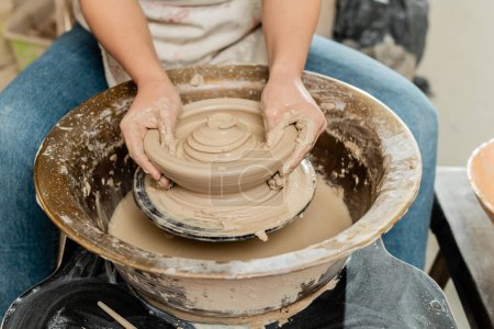 Téléchargez les photos : Vue recadrée de l'artisan femelle floue dans le tablier faisant la forme d'argile humide sur la roue de poterie filante tout en travaillant dans un atelier de céramique, concept de fabrication de poterie qualifiée - en image libre de droit