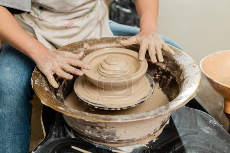 Cropped view of young craftswoman in apron making shape of clay on spinning pottery wheel in blurred ceramic workshop at background, skilled pottery making concept