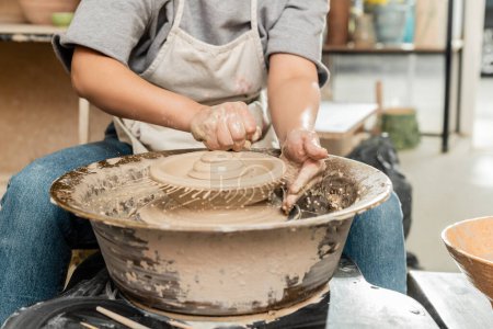 Téléchargez les photos : Vue recadrée du jeune artisan femelle dans le tablier versant de l'eau de l'éponge sur l'argile sur la roue de poterie en rotation près du bol dans un studio d'art flou, processus de création de poterie - en image libre de droit