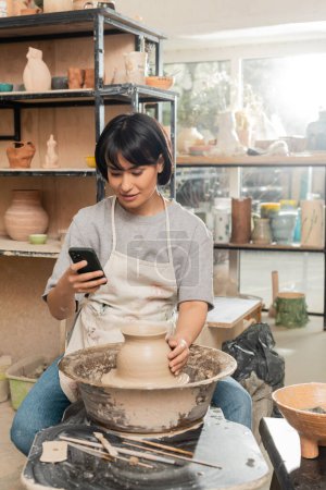 Sonriente asiática morena artesana en delantal usando teléfono inteligente mientras trabaja con arcilla en la rueda de cerámica cerca de herramientas de madera y tazón en taller de cerámica, proceso de creación de cerámica