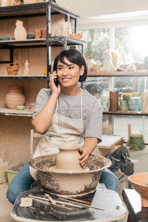 Fröhliche junge asiatische Kunsthandwerkerin in Schürze, die mit dem Smartphone spricht und neben Ton auf Töpferscheibe und Holzwerkzeug in der Keramikwerkstatt sitzt, Töpferei-Entstehungsprozess