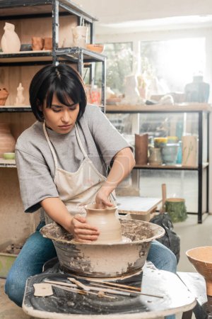 Junge brünette asiatische Kunsthandwerkerin in Schürze Form Ton Vase während der Arbeit an Töpferscheibe in der Nähe von Holzwerkzeugen und Schale in verschwommener Keramik-Werkstatt, Keramik-Entstehungsprozess
