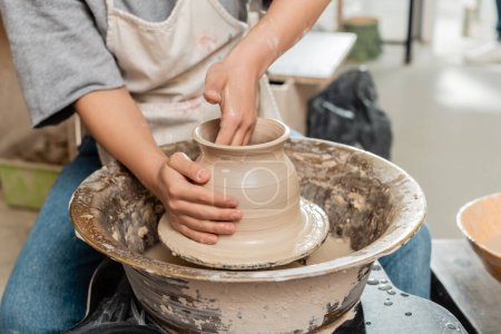 Vue recadrée du jeune potier femelle dans un vase en argile de moulage de tablier et travaillant avec la roue de poterie tournante dans un atelier de céramique floue en arrière-plan, processus de création de poterie