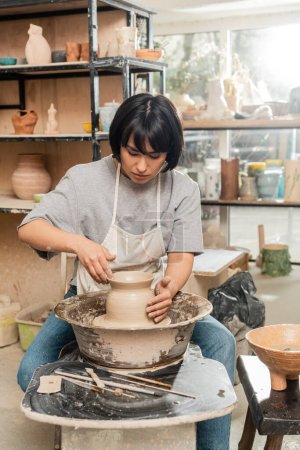 Young asian brunette female artisan in apron and workwear making clay vase and working on spinning pottery wheel in blurred ceramic workshop at background, pottery creation process