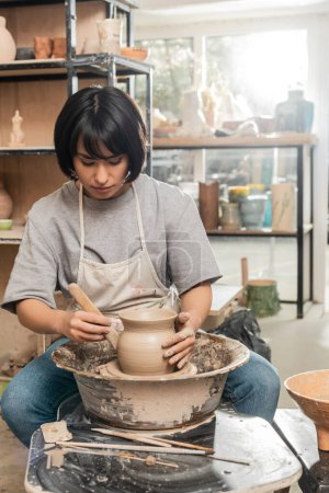 Joven morena asiática alfarera en delantal sosteniendo herramienta de madera cerca de arcilla mientras trabaja con la rueda de cerámica giratoria en taller de cerámica borrosa, proceso de creación de cerámica