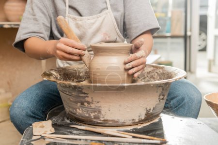 Vista recortada de la joven artesana en delantal haciendo forma de jarrón de arcilla con herramienta de madera en la rueda de cerámica giratoria en taller de cerámica borrosa en el fondo, proceso de creación de cerámica