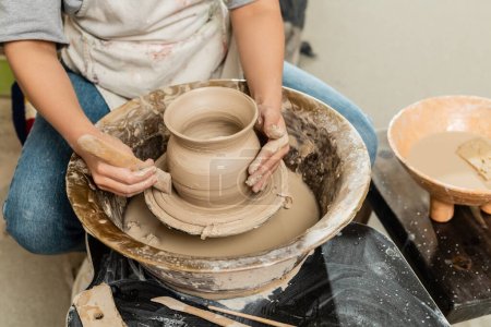Téléchargez les photos : Vue recadrée du jeune potier femelle en tablier faisant forme de vase en argile avec outil en bois sur la roue de poterie près du bol avec de l'eau et de l'éponge en atelier d'art, façonnage et formage de l'argile processus - en image libre de droit
