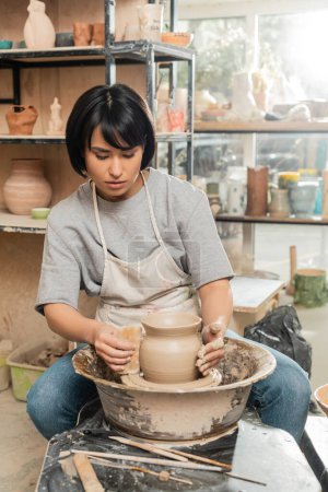 Young brunette asian craftswoman in apron holding scraper near clay vase on spinning pottery wheel near wooden tools on table in blurred ceramic workshop, clay shaping and forming process