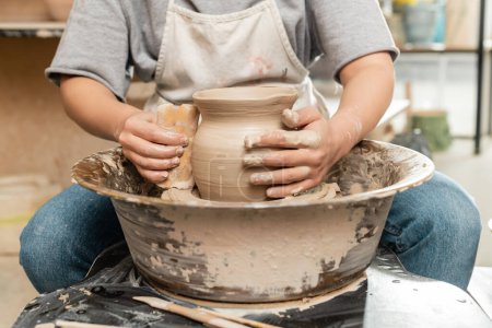 Vue recadrée du jeune potier femelle en tablier faisant forme de vase en argile avec grattoir en bois sur la roue de poterie tournante en atelier d'art, façonnage et formage de l'argile