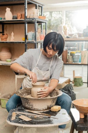 Junge brünette asiatische Handwerkerin in Schürze, die die Form einer Tonvase herstellt, während sie mit der Töpferscheibe in der Nähe von Schwamm und Holzwerkzeugen in der Keramikwerkstatt arbeitet, Formen und Formen von Ton