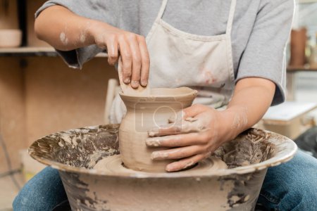 Cropped view of young female artisan in apron making shave of clay vase with wooden tool while working with spinning pottery wheel in ceramic workshop, clay shaping and forming process