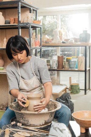 Joven mujer asiática artesana en delantal haciendo forma de jarrón de arcilla con esponja húmeda y trabajando con rueda de cerámica giratoria en taller de cerámica borrosa, moldeado y proceso de formación de arcilla