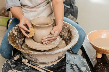 Téléchargez les photos : Vue recadrée du potier femelle en tablier en forme de vase en argile avec éponge vétérinaire près des outils en bois et de la roue de poterie en atelier de céramique, façonnage et formage de l'argile - en image libre de droit