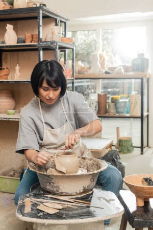 Brunette asian female artisan in apron cutting wet clay on spinning pottery wheel while working near wooden tools and bowl in blurred ceramic workshop, clay shaping and forming process