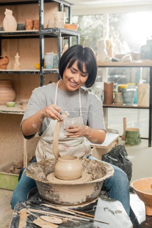 Cheerful young brunette asian artisan in apron holing clay while making vase on pottery wheel near tools and bowl in blurred ceramic workshop, artisanal pottery production and process