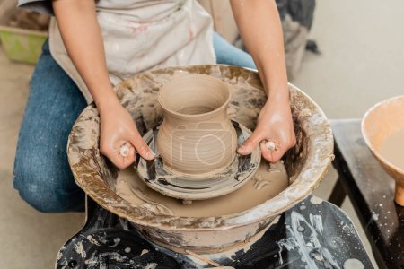 Cropped view of young female artisan in apron cutting clay vase on spinning pottery wheel near bowl with water at background in ceramic workshop, artisanal pottery production and process