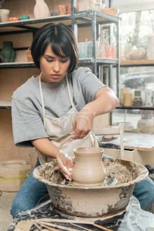 Junge brünette asiatische Kunsthandwerkerin in der Werkstatt, die Tonkrüge herstellt, während sie am Spinnrad in der Nähe von verschwommenen Holzwerkzeugen in der Keramikwerkstatt, der handwerklichen Töpferei-Produktion und -Verarbeitung arbeitet
