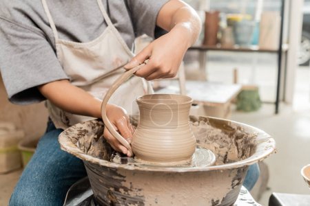 Vista recortada de alfarera joven en delantal haciendo jarra de arcilla mientras se trabaja con rueda de cerámica en taller de cerámica borrosa en el fondo, producción y proceso de cerámica artesanal