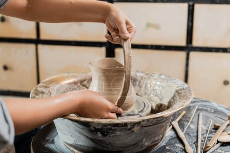 Vista recortada de la joven ceramista haciendo jarra de arcilla y trabajando con rueda de cerámica cerca de herramientas de madera en taller de arte borroso, producción y proceso de cerámica artesanal