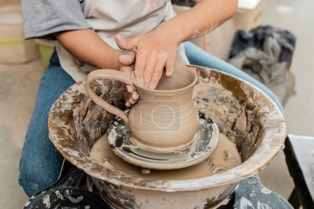 Vista recortada de la joven artista femenina en delantal creando jarra de arcilla en la rueda de cerámica en la mesa mientras trabaja en taller de cerámica, producción y proceso de cerámica artesanal