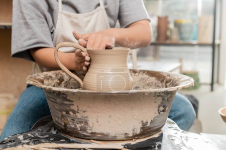 Ausgeschnittene Ansicht einer jungen Kunsthandwerkerin in Schürze, die Tonkrüge herstellt, während sie mit der Töpferscheibe auf dem Tisch in einer verschwommenen Keramikwerkstatt arbeitet, handwerkliche Töpferei-Produktion und -Prozess