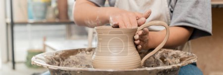 Photo for Cropped view of blurred young female potter in apron and workwear making clay jug while working with pottery wheel in art studio at background, clay shaping technique and process, banner - Royalty Free Image
