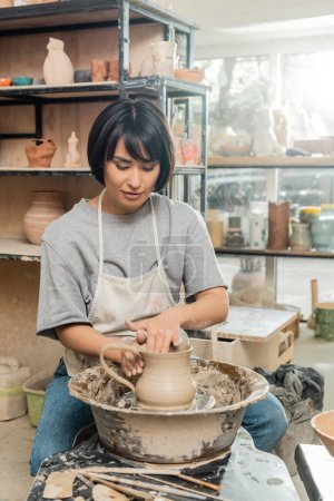 Junge asiatische Künstlerin in Schürze und Arbeitskleidung, die Tonkrug auf Töpferscheibe in der Nähe von Holzwerkzeugen auf dem Tisch in verschwommener Keramikwerkstatt im Hintergrund, Tonformtechnik und -prozess