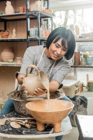Smiling brunette asian craftswoman in apron pouring water from clay jug in bowl near pottery wheel and wooden tools on table in blurred art studio, clay shaping technique and process