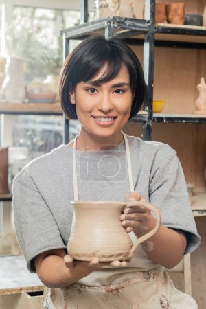 Porträt einer jungen brünetten asiatischen Kunsthandwerkerin in Schürze und Arbeitskleidung mit Tonkrug und Blick auf die Kamera in einer unscharfen Keramikwerkstatt im Hintergrund, Technik und Prozess der Tonformung