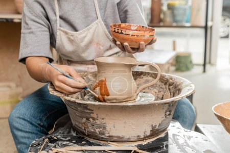 Photo for Cropped view of young female artist in apron painting on clay jug on pottery wheel near wooden tools n table in blurred ceramic workshop, clay shaping technique and process - Royalty Free Image