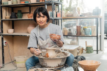 Cheerful young asian craftswoman in apron painting clay jug and looking at camera near pottery wheel and wooden tools in art workshop at background, clay shaping technique and process