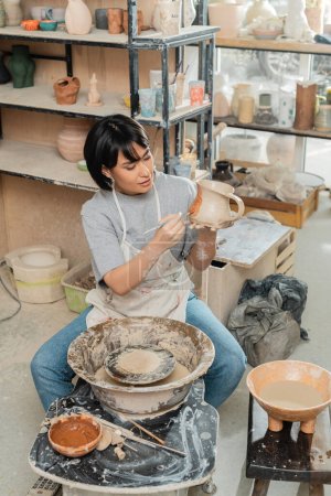 High angle view of young asian female artisan in apron painting on clay jug near pottery wheel, wooden tools and bowl with water in ceramic workshop, clay shaping technique and process