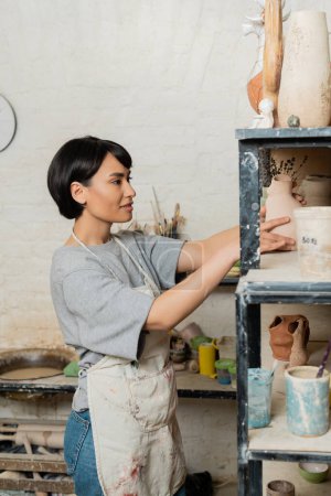Lächelnde junge asiatische Kunsthandwerkerin in Schürze stellt Keramik-Skulptur auf Regal, während sie im verschwommenen Kunstworkshop im Hintergrund steht, Technik und Prozess der Tonformung