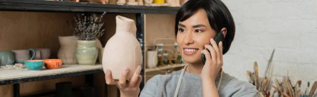 Smiling young brunette asian craftswoman talking on smartphone and holding ceramic sculpture and standing near rack in ceramic workshop at background, pottery studio scene, banner 
