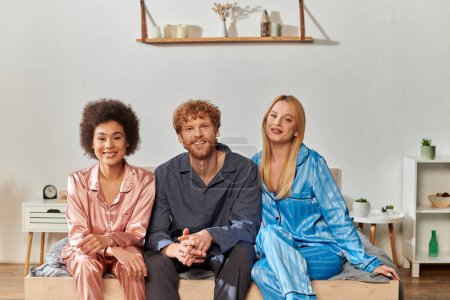 Photo for Open relationship, polygamy, understanding, three adults, redhead man and multicultural women in pajamas sitting on bed at home, cultural diversity, acceptance, bisexual - Royalty Free Image