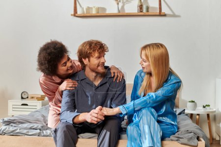 Photo for Open relationship concept, three adults, redhead man holding hands with blonde woman, multicultural people in pajamas sitting on bed at home, cultural diversity, acceptance, bisexual - Royalty Free Image