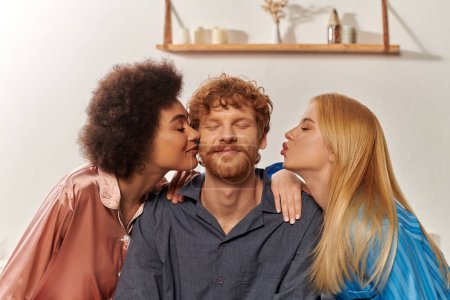 Polygamie-Konzept, offene Beziehung, Porträt von drei Erwachsenen, multikulturelle Frauen küssen glücklichen Rotschopf, polyamouröse Familie im Pyjama, kulturelle Vielfalt, Akzeptanz, bisexuell, polyamory 