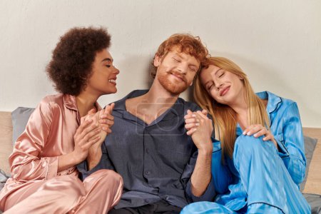 offene Beziehung, Polygamie, glückliche drei Erwachsene, rothaariger Mann und multikulturelle Frauen im Schlafanzug, Händchenhalten im Bett, kulturelle Vielfalt, Akzeptanz, bisexuell, moderne Familie 