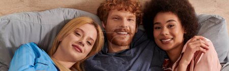 polygamy, alternative relationships, three adults, positive redhead man and multicultural women lying under blanket together, cultural diversity, acceptance, bisexual, open relationship, banner 