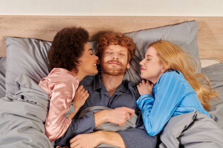 Polyamory-Konzept, drei Erwachsene, glücklicher Mann und gemischtrassige Frauen im Schlafanzug, morgens gemeinsam aufwachen, unter Decke, Schlafzimmer, kulturelle Vielfalt, bisexuell, offene Beziehung, Polygamie, Draufsicht