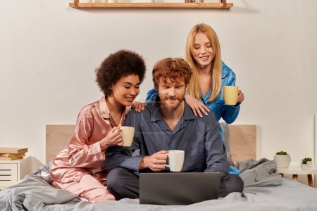 polygamy, multicultural women and redhead man in pajamas watching movie on laptop, holding cups of coffee in bedroom, cultural diversity, acceptance, bisexual, open relationship 